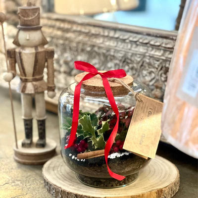 Terrarium in vetro con piante e fiocco rosso su base in legno, accanto a un soldatino natalizio